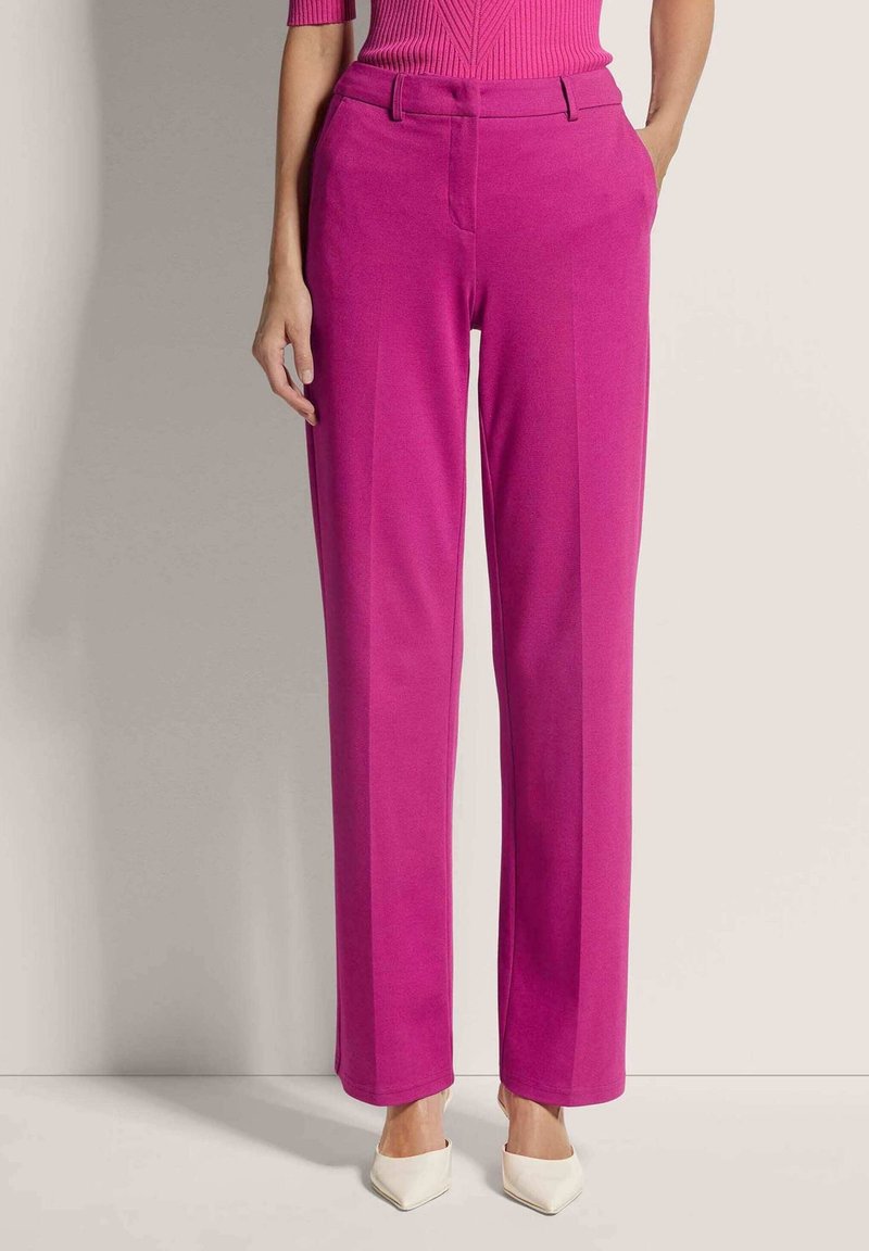 Femme portant un pantalon rose vif ajusté avec un pli sur le devant, associé à des talons blancs à bout pointu, debout devant un fond uni.