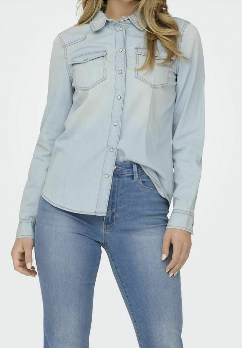 Femme portant une chemise en denim bleu clair et un jean bleu, avec la chemise partiellement rentrée, debout devant un fond blanc.