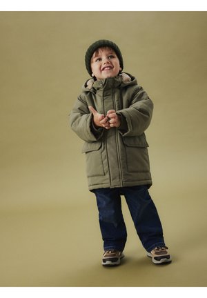 Niño sonriente que lleva un gorro de punto verde, una chaqueta acolchada verde oliva, unos jeans azules y deportivas, de pie sobre un fondo beige liso.