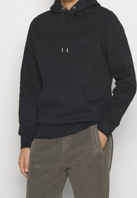Uomo che indossa una felpa nera con cappuccio e tasca frontale, pantaloni grigi, con una mano in tasca di fronte a uno sfondo semplice.