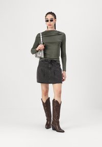 Maglia verde a maniche lunghe, gonna di jeans nera a vita alta e stivali da cowboy marroni con dettagli ricamati. Piccola borsa argento e occhiali da sole.