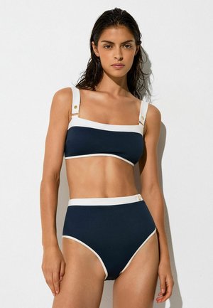 Kvinde med vådt hår, iført en navyblå bikini med høj talje og hvid kant, stående foran en ensfarvet hvid baggrund.