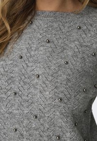 Szary sweter z splotem w jodełkę i dekoracyjnymi metalowymi ćwiekami. Materiał wydaje się być miękki i dziany, z dekoltem typu crew neck.