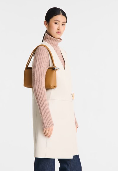 Tasche aus braunem Leder mit strukturierter Form und kurzen Griffen, getragen über einem hellcremefarbenen ärmellosen Kleid und einem pinken, gerippten Rollkragenpullover.