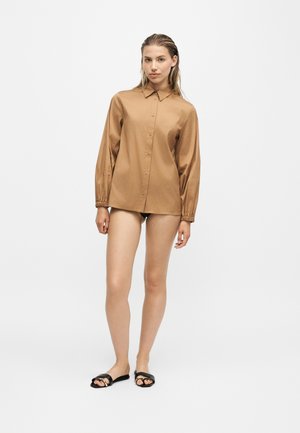 Femme debout portant une chemise marron ample boutonnée, des sous-vêtements noirs et des sandales noires sur un fond blanc uni.