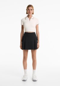 Egy nő áll, testhez simuló, fehér, rövid ujjú polo crop topot, fekete, plisszírozott mini szoknyát, fehér zoknit és fehér sportcipőt visel.