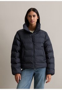 Doudoune noire avec un col haut, fermeture éclair et texture matelassée. Le modèle porte un jean clair et un pull crème en dessous.