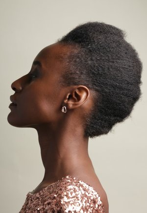Profielweergave van een persoon met natuurlijk, textuurvol zwart haar dat omhoog gestyled is. Draagt ronde, roségouden oorbellen en een pailletten top in roségoud.