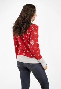 Red holiday sweater with white snowflake and polka dot patterns. Features a ribbed white hem and cuffs. Worn over dark jeans.