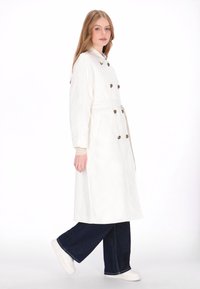 Witte trenchcoat met een dubbelgeknoopte uitstraling, grote knopen en een ceintuur. Gedragen over donkerblauwe wijde jeans en witte sneakers.