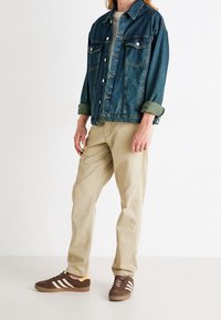 Spijkerjack met een vervaagde blauwe afwerking, knoopsluiting aan de voorkant, twee borstzakken; gecombineerd met beige broek en bruine sneakers met witte strepen.