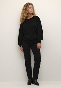 Sort sweater med afslappet pasform og sænkede skuldre, parret med sorte slim-fit bukser. Modellen har spidse sorte ankelstøvler på.