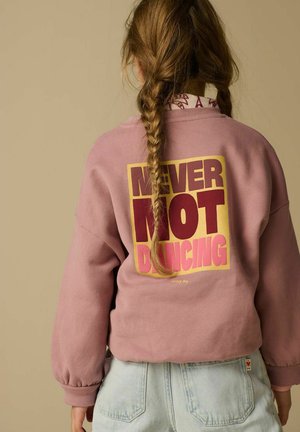 Enfant aux longs cheveux tressés portant un sweat-shirt rose avec le texte "NEVER NOT DANCING" en lettres majuscules au dos.