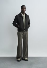 Mies seisoo käsineen taskuissa, yllään tummanvärinen vetoketjullinen takki, kuvioitu paita, leveälahkeiset harmaat housut ja kaksiväriset kengät harmaalla lattialla.