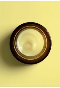 Bernsteinfarbener Glasbehälter mit einem goldenen Rand, gefüllt mit einer glatten, blassgelben Creme. Die Oberfläche hat ein leicht wirbelndes Muster. Der Hintergrund ist ein einheitliches Gelb.