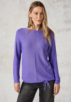 Haut à manches longues violet en tissu texturé, encolure ronde et ourlet à cordon de serrage. Porté avec un jean foncé, sur un fond neutre.