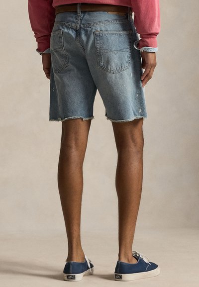 Άνδρας που φοράει ανοιχτόχρωμα τζιν σορτς με ξεφτισμένα τελειώματα και μπλε πάνινα αθλητικά παπούτσια, στέκεται με την πλάτη να φαίνεται σε απλό φόντο.