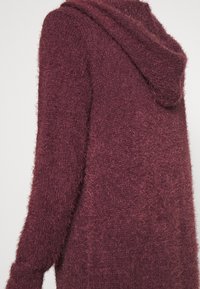 Cardigan lavorato in bordeaux con una consistenza morbida e soffice e cappuccio. Presenta maniche lunghe e una vestibilità rilassata, progettato per comfort e calore.