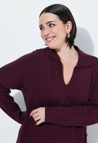 Sweter dzianinowy w głębokim burgundzie, z dekoltem w kształcie litery V, długimi rękawami i ściągaczami na mankietach. Miękka tekstura, luźny krój i subtelne szwy na ramionach.