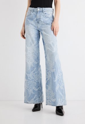 Jasnoniebieskie dżinsy z szerokimi nogawkami ozdobione białą kwiatową haftem, noszone z czarnymi kozakami na obcasie i czarną bluzką.