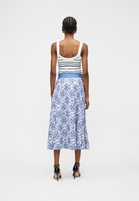 Femme aux cheveux tressés portant un débardeur blanc à rayures bleu marine, une jupe midi bleue à motifs et des sandales noires à talons hauts, debout, dos tourné vers l'avant.