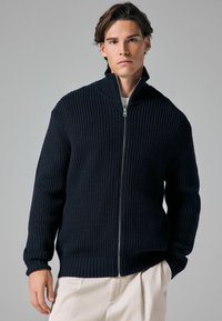 Marine gebreide zip-up jack met een hoge kraag, geribbelde textuur en lange mouwen, gecombineerd met lichtgekleurde broeken.