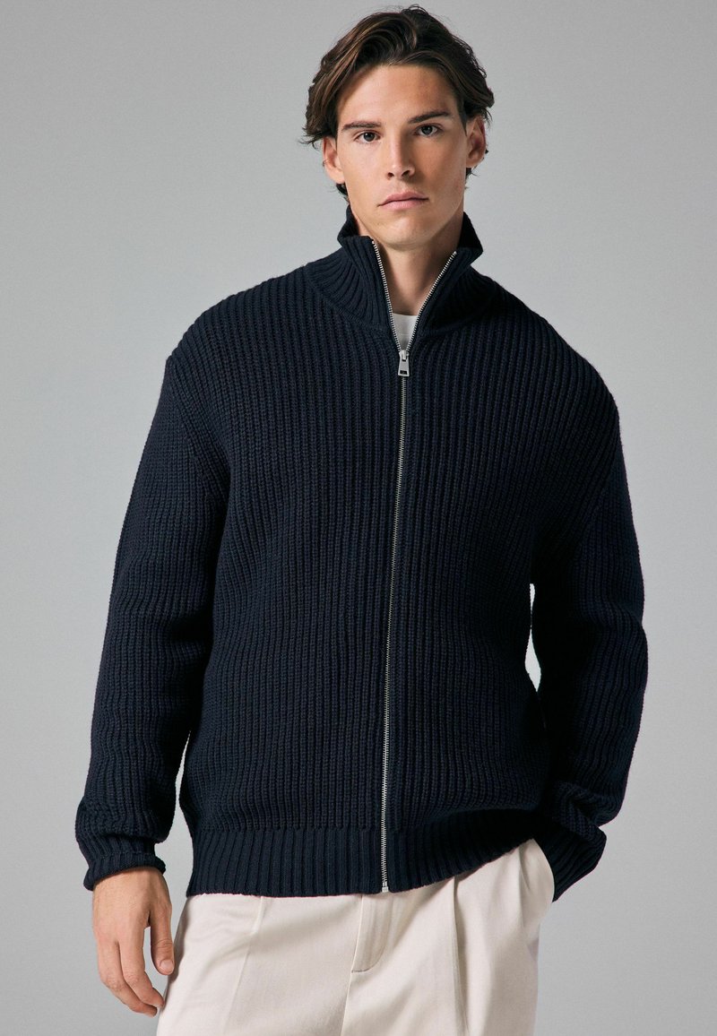 Marine gebreide zip-up jack met een hoge kraag, geribbelde textuur en lange mouwen, gecombineerd met lichtgekleurde broeken.