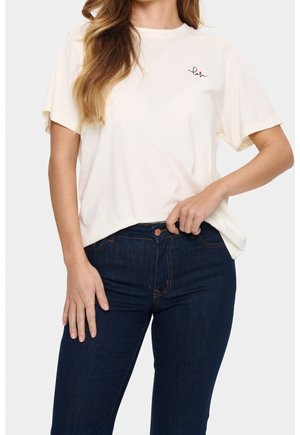 Woman wearing a white short-sleeve t-shirt with “love” text and a small red heart on the left chest, paired with dark blue jeans.