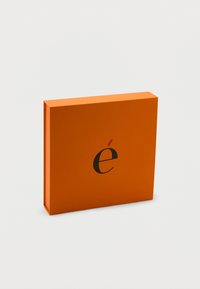 Ορθογώνιο πορτοκαλί κουτί με ένα μαύρο μικρό γράμμα «é» και κόκκινη έμφαση στο κέντρο της μπροστινής όψης, τοποθετημένο μπροστά σε απλό λευκό φόντο.