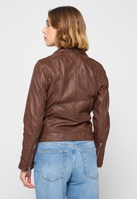 Brązowa skórzana kurtka o dopasowanym kroju, z wyraźnie zarysowanymi ramionami i zamkami na rękawach. Połączenie z jasnoniebieskimi dżinsami tworzy kontrast.