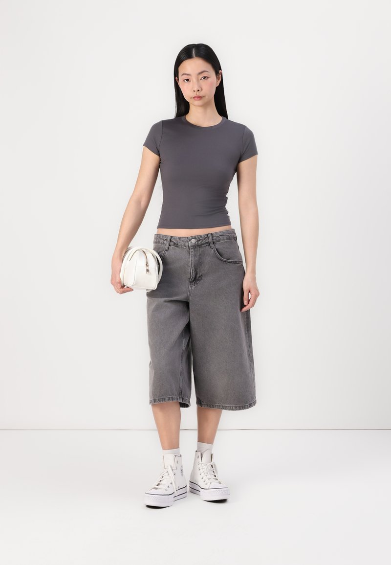 Grau gefittetes T-Shirt kombiniert mit lockeren grauen Denim-Culottes. Das Model hält eine kleine weiße Handtasche und trägt weiße Sneakers mit hohem Schnitt.