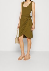 Robe portefeuille vert olive avec encolure dégagée et détail de lien sur le côté, associée à des sandales à talons blocs nude et un petit sac à main clair.