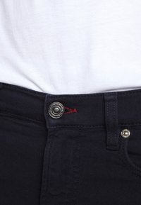 Mörkblå jeans med en metallknapp med ingraverade detaljer och röd söm på midjebandet. Bomullstyg med en mjuk och jämn textur.