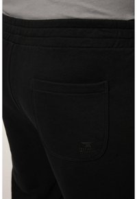 Czarne dresy mają elastyczny pas, tylną kieszeń i mały detal z logo. Materiał wydaje się gładki i miękki.