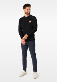 Maglione nero con polsini e orlo a coste, caratterizzato da un logo grafico multicolore sul petto. Indossato con pantaloni a fantasia blu scuro e sneakers bianche.