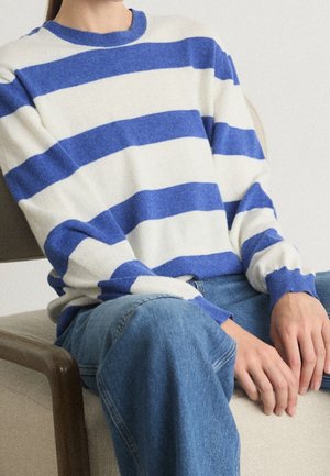 Person sitting on beige chair wearing blue and white horizontally striped sweater and blue jeans, hands resting on lap.
