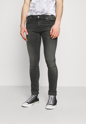 Mann trägt schwarze Slim-Fit-Jeans, an den Knöcheln hochgekrempelt, und schwarze High-Top-Sneaker mit weißen Schnürsenkeln, steht vor einem schlichten hellen Hintergrund.