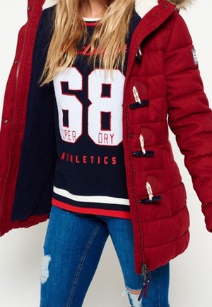 Personne portant un manteau matelassé rouge avec des boutons à bascule ouvert sur un sweat bleu marine "68 Superdry Athletics" et un jean bleu déchiré.