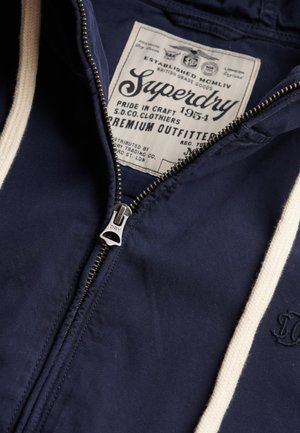 Marineblauwe hoodie met crèmekleurige trekkoorden, zichtbare metalen ritssluiting en een groot Superdry-merklabel aan de binnenkant van de kraag met gedetailleerde tekst.