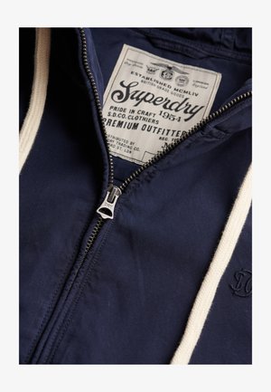 Marineblauwe hoodie met crèmekleurige trekkoorden, zichtbare metalen ritssluiting en een groot Superdry-merklabel aan de binnenkant van de kraag met gedetailleerde tekst.