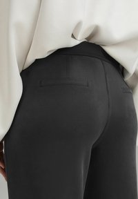 Gros plan d’une personne portant un pantalon foncé ajusté avec des poches passepoilées à l’arrière et un chemisier beige clair rentré.