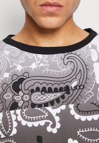 Košile s krátkým rukávem s vzorem v šedé, černé a bílé barvě s motivem paisley. Má černý žebrovaný límec a vyšívané jméno značky.