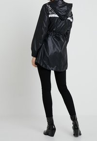 Černá kapucová bunda s lesklým povrchem, krajkovými detaily na ramenou, stahovací pas a elastické manžety. V kombinaci s černými legínami a botami na vysokém podpatku.
