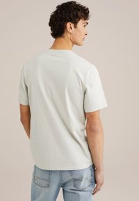 Světle zelené bavlněné tričko s kulatým výstřihem a krátkými rukávy, zobrazené ze zadní strany. V kombinaci s modrými džíny.