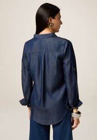 Cămașă din denim de culoare albastru închis, cu mâneci lungi, buzunar pe piept și manșete cu butoane. Are un tiv ușor curbat și o croială lejeră.