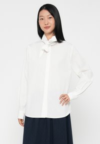 Camicia bianca a maniche lunghe con colletto a punta e sciarpa annodata al collo, con una consistenza liscia e una vestibilità dritta.