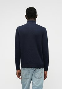 Maglione blu navy con texture e motivo in rilievo, colletto e polsini a costine, abbinato a jeans di denim blu chiaro. Vista dalla schiena.