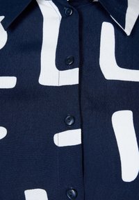 Chemise bleu marine avec de grandes formes abstraites blanches et des boutons noirs, montrant les détails du col et de la patte de boutonnage.