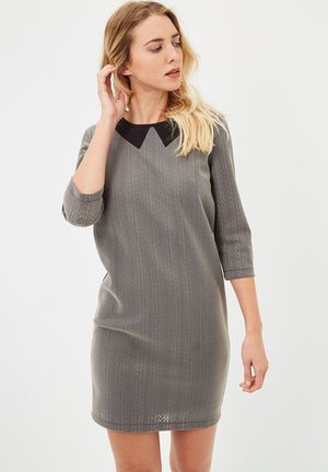 Joven mujer con cabello rubio, vestida con un vestido gris texturizado hasta la rodilla, con mangas tres cuartos y un detalle de cuello negro, mirando hacia la izquierda.