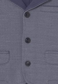 Granatowa marynarka w wzór jodełki z zapięciem na dwa guziki, kieszeniami naszytymi oraz teksturowanym materiałem z kontrastowym przeszyciem przy klapie.